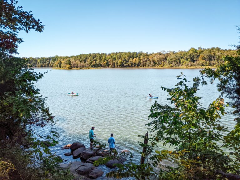 Two people bank fishing on a rock at Latta Nature Preserve. Two kaykers paddle in the distance.