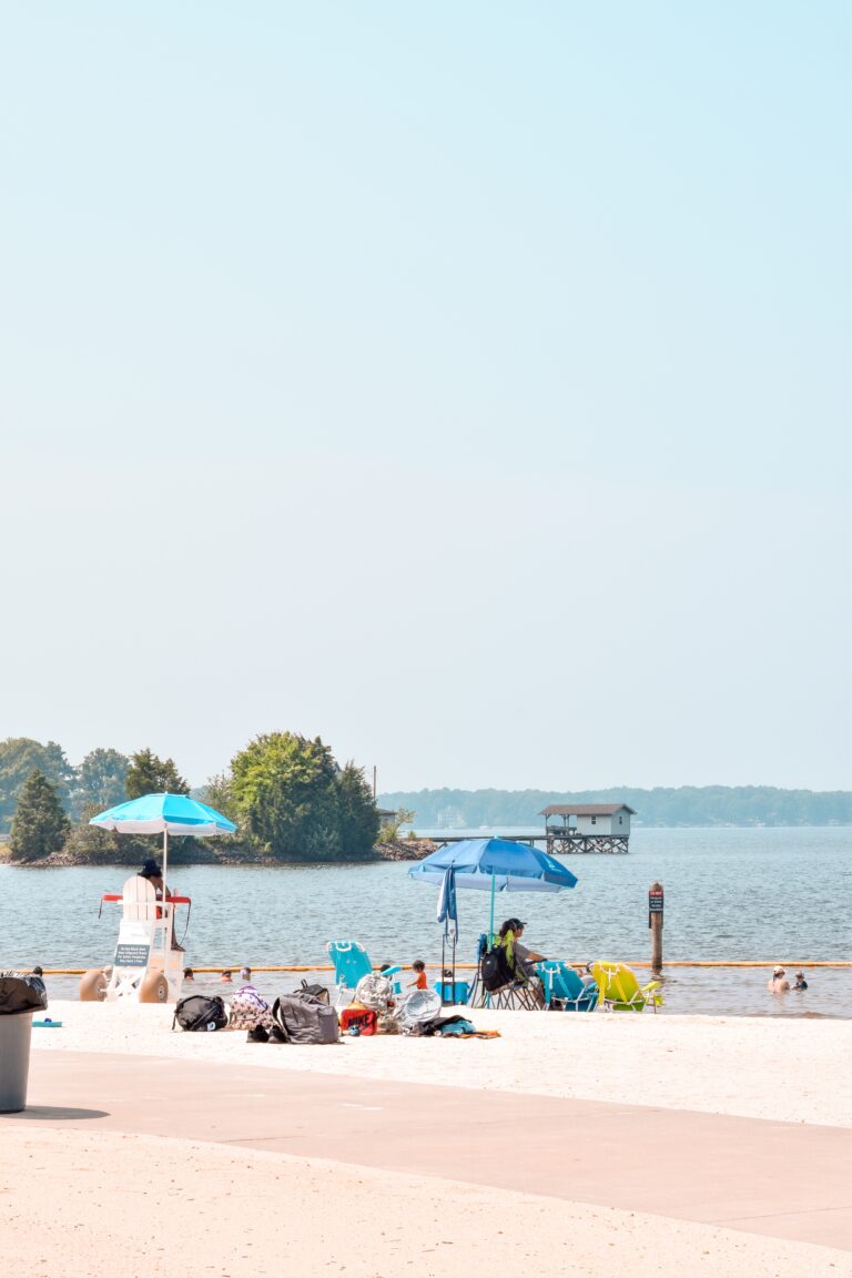 People relax on the beach near Jetton Park at Ramsey Creek underneath blue umbrellas with the lake in the background.
